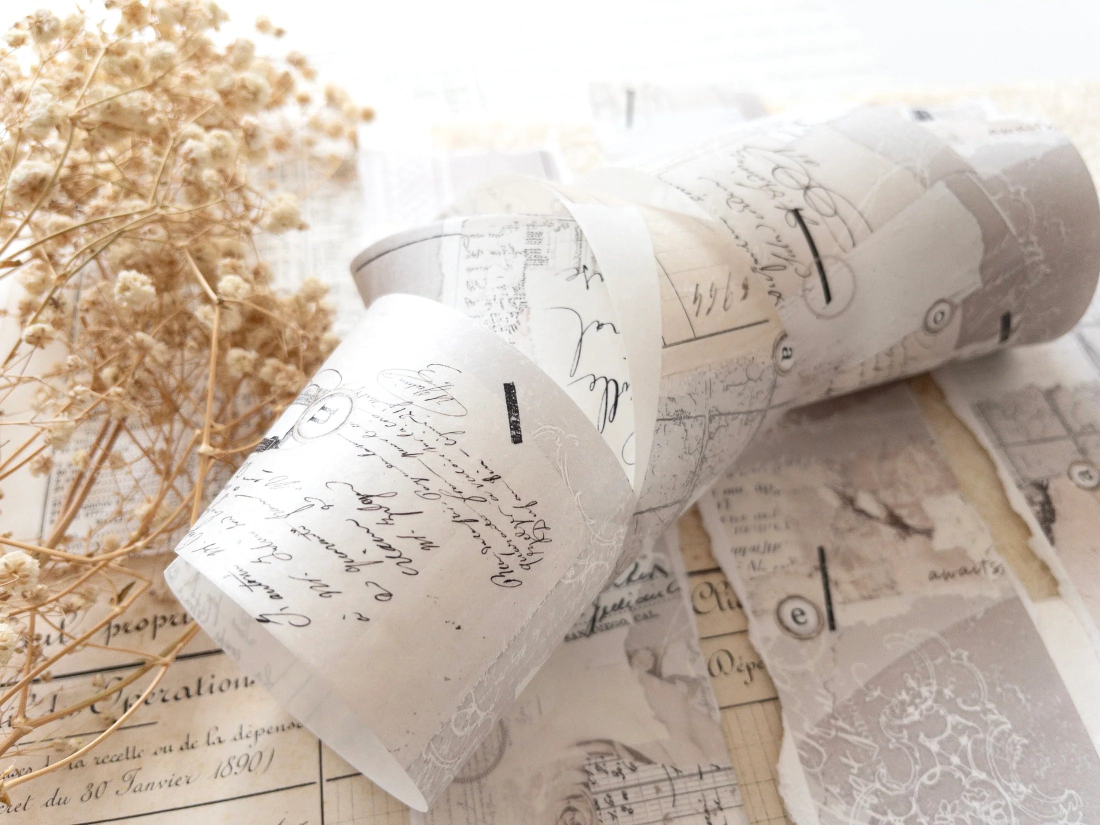 Decorative washi tape with vintage designs rolled up on a white surface with dried flowers.