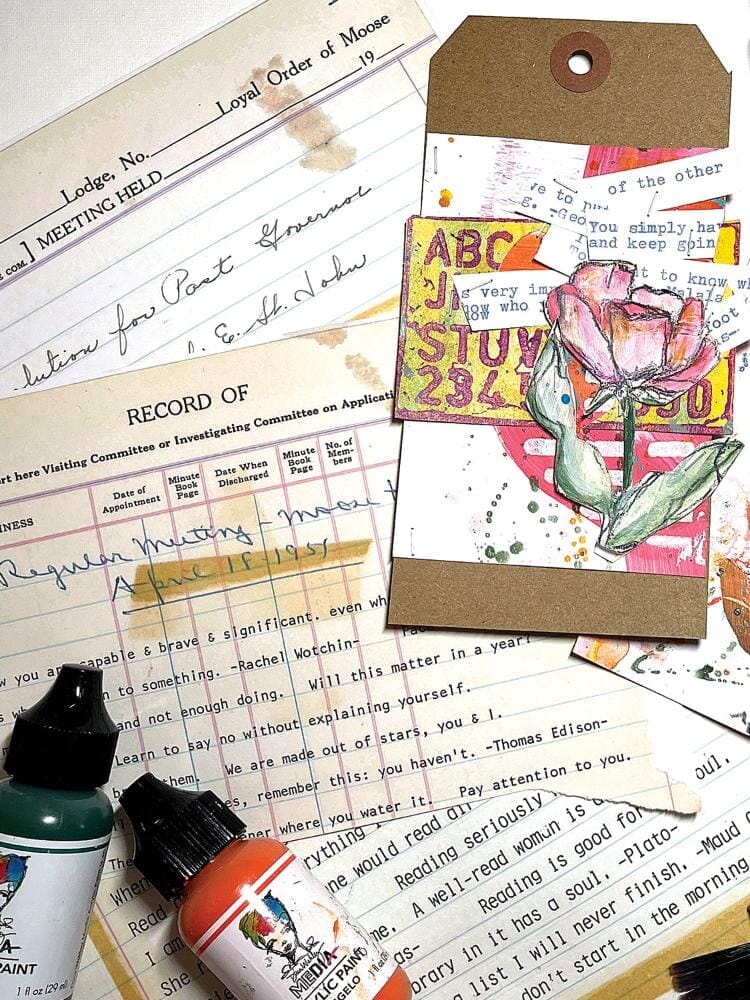 A styled flat-lay of mixed-media art supplies and scrapbook papers, featuring a kraft paper tag with a pink rose illustration, paint bottles, and vintage-style printed papers.
