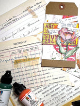 A styled flat-lay of mixed-media art supplies and scrapbook papers, featuring a kraft paper tag with a pink rose illustration, paint bottles, and vintage-style printed papers.