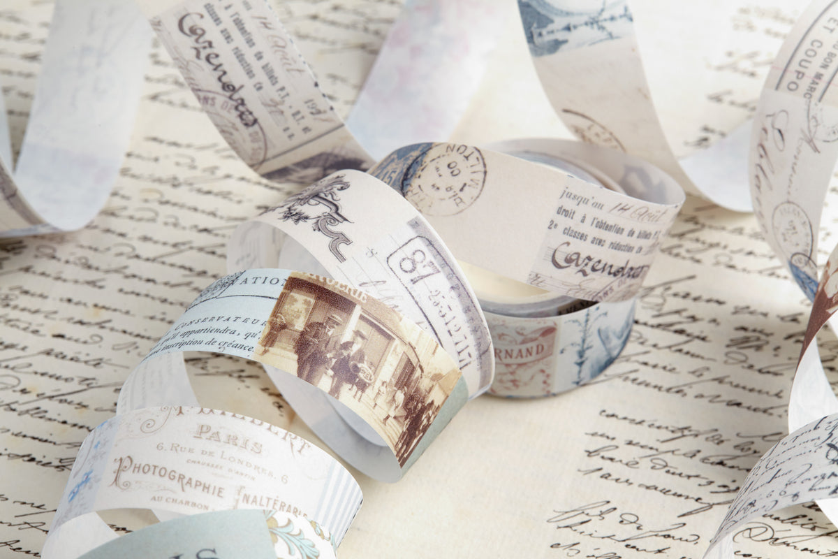Vintage-style washi tape roll printed with old postmarks, stamps, and script, arranged on top of a sheet of cursive handwriting.