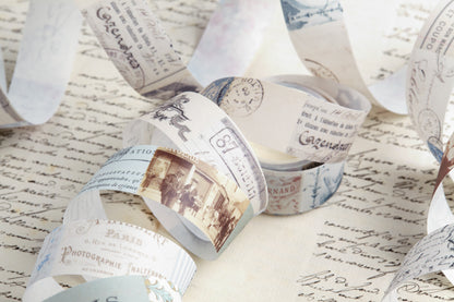 Vintage-style washi tape roll printed with old postmarks, stamps, and script, arranged on top of a sheet of cursive handwriting.