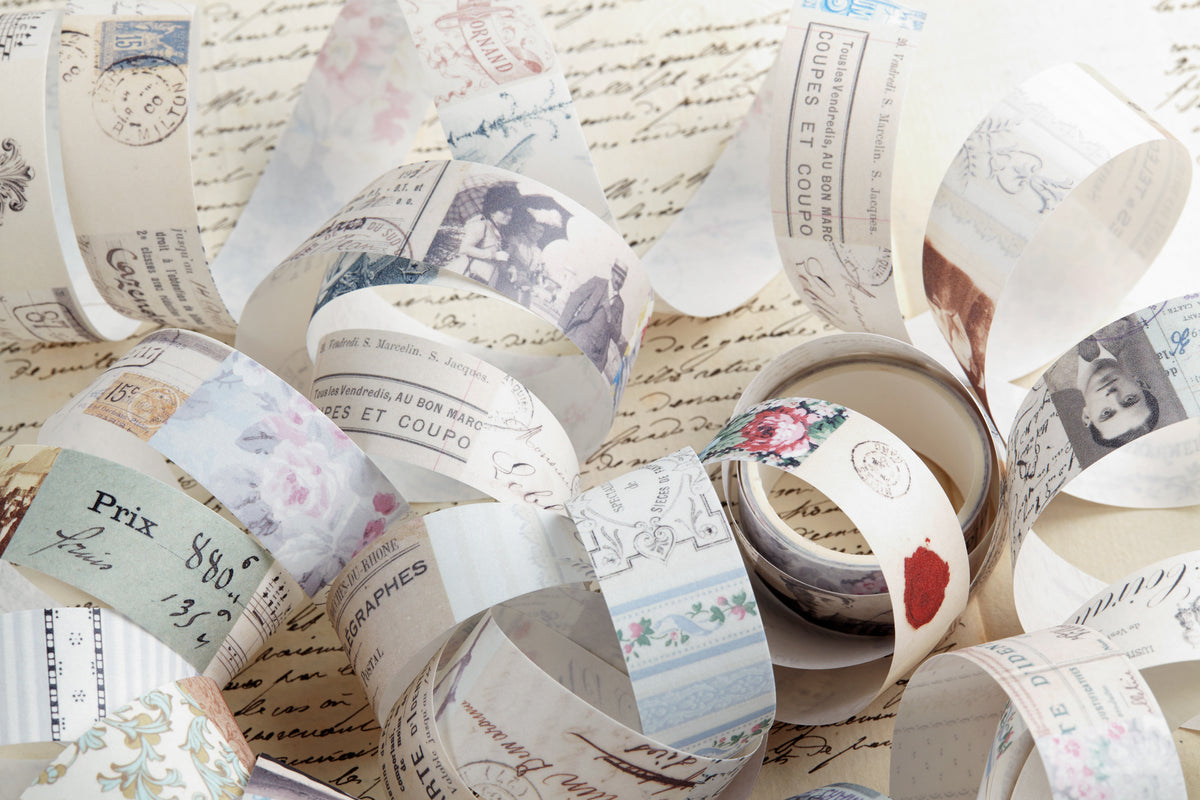A pile of vintage-style decorative washi tape with French script, postmarks, floral prints, and illustrated portraits, arranged on a background of cursive handwriting paper.