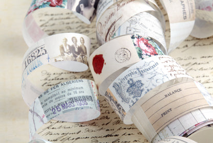 A close-up of a washi tape rolls with vintage-style printed designs, including script, numbers, floral motifs, and illustrated figures, arranged on top of a parchment-like background with cursive writing.