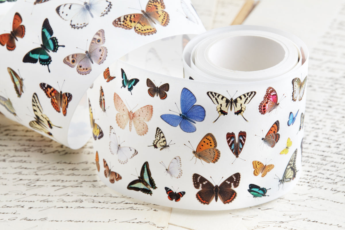 A roll of pre-cut washi tape featuring a colorful vintage butterflies on a white background, styled on top of antique handwritten book pages.