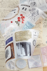 PET tape with pre-cut vintage-style decorative stickers such as illustrated labels, postage stamps, numbers, and antique typography, laid over a background of old handwritten letters and papers.