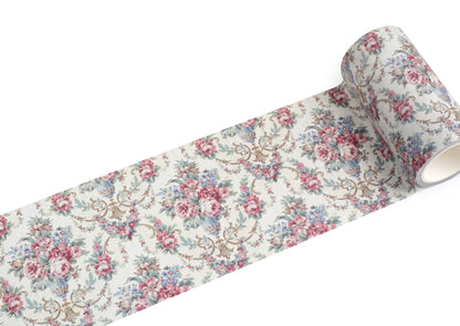 A roll of extra wide vintage-style floral washi tape with a repeating pattern of pink and blue roses, green leaves, and tan scrollwork on a warm off-white background, shown unrolled against a plain white studio background.