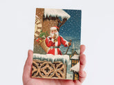 A hand holding a rectangular Christmas postcard featuring Santa Claus in a snowy village scene, with snow-covered rooftops, a large clock, and falling snow in the background.