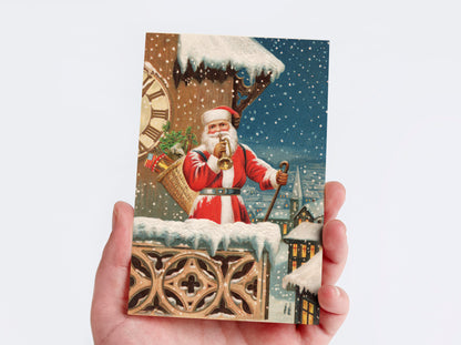 A hand holding a rectangular Christmas postcard featuring Santa Claus in a snowy village scene, with snow-covered rooftops, a large clock, and falling snow in the background.