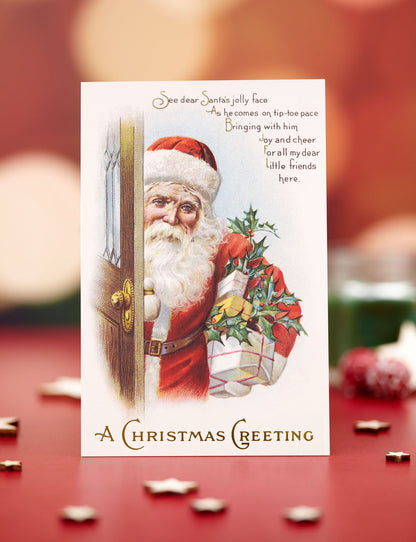 A vintage-style Christmas greeting card featuring an illustration of Santa Claus peeking from behind a door with a message, displayed on a red surface with blurred holiday lights in the background.