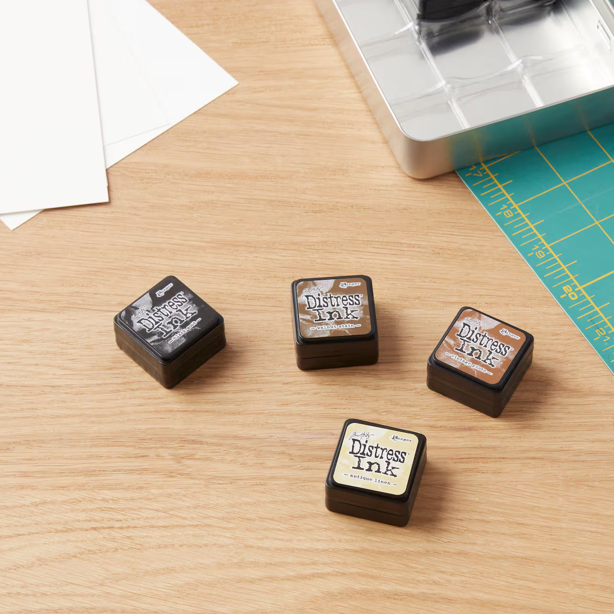 Four Distress Ink stamp pads in different colors arranged on a light wood desk with crafting tools nearby, including white paper, a metal tray, and a green cutting mat.