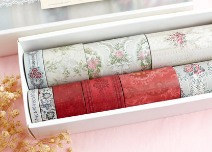 A white gift box containing multiple rolls of decorative washi tape with vintage floral and damask patterns in red, green, pink, and blue, arranged on a soft pink background with dried flowers.