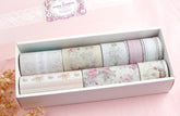 A white rectangular box containing nine rolls of decorative washi tape with pastel floral and striped patterns, set on a soft pink background with a small dried flower sprig.