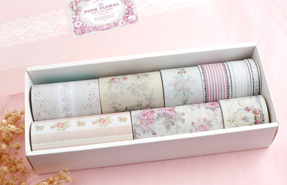 A white rectangular box containing nine rolls of decorative washi tape with pastel floral and striped patterns, set on a soft pink background with a small dried flower sprig.