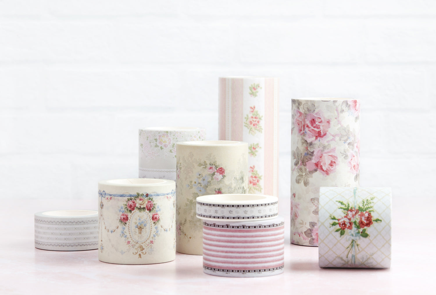 A collection of decorative paper washi tapes with floral and striped patterns, arranged on a light pink surface against a white brick wall background.