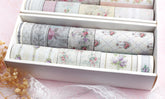 A white box filled with rolls of decorative washi tapes featuring vintage floral and damask patterns in soft pastel colors, arranged on a pink surface with lace and dried flowers.