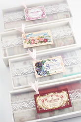 A line of four clear plastic lid boxes with white lace trim and vintage-style paper labels, shown on a white background.