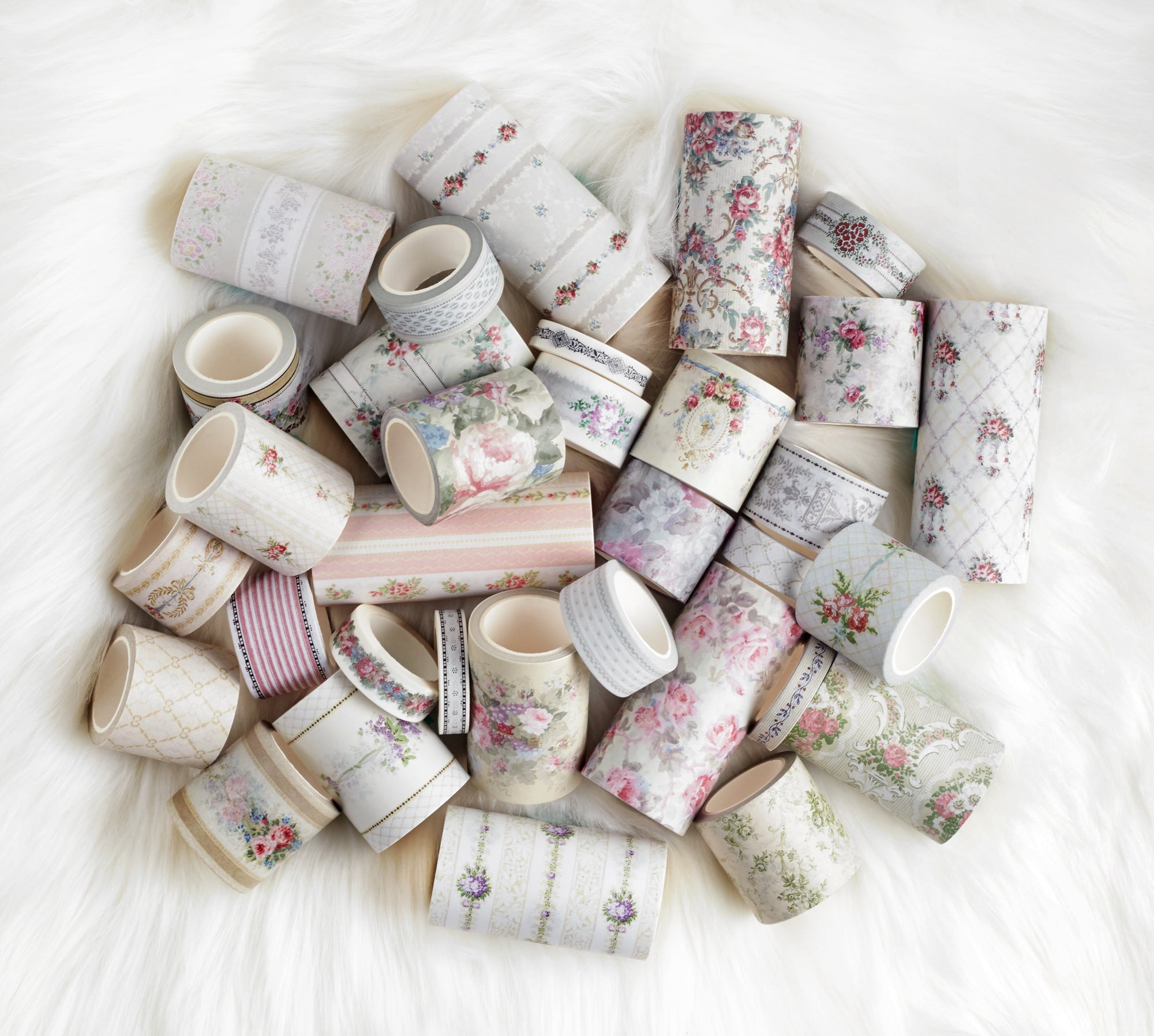 A pile of decorative washi tapes with various floral, geometric, and striped patterns in soft pastel colors, arranged on a white faux fur surface.